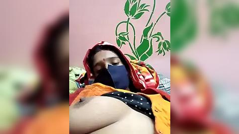 Media: A blurry video shows a woman with medium skin tone, wearing a red and yellow sari, lying on a bed. She has a blue face mask and her breasts are exposed. The background features green floral wallpaper and a red and green patterned bedspread.