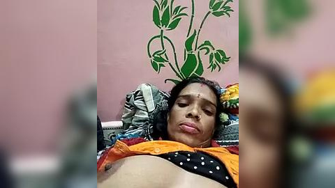 Media: Video of a woman with medium brown skin, lying on a bed with a colorful blanket. She has a green plant on her head and is wearing a black dress with white polka dots and orange fabric. The background is a pink wall with a decorative pattern.