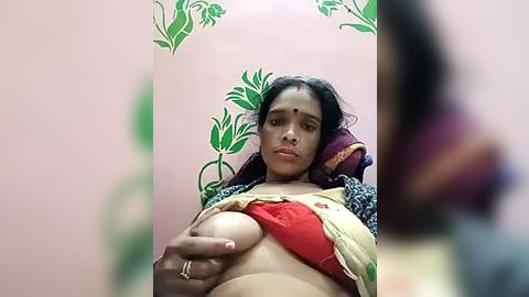 Media: Video of a South Asian woman with medium skin tone, dark hair, and a red bra, holding a breast, in a room with green floral wallpaper.