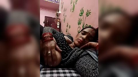 Media: Video of an Indian woman with medium skin tone, lying on a bed, wearing a black polka-dot dress, her legs bound with red rope, in a room with pink walls and green plant decoration.