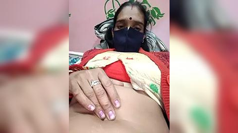 Media: Video of a woman with light brown skin, wearing a red and white patterned top, a blue mask, and a ring on her finger, lying on a bed.