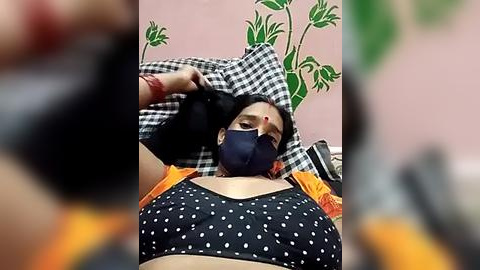 Media: Video of a woman with medium skin tone and dark hair, wearing a black face mask, lying on a bed with a black polka-dot bra and orange shirt. Pink wall with green leaf stickers in the background.