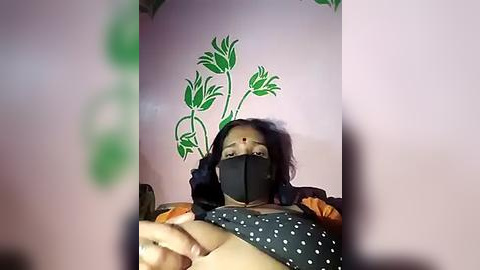 Media: Video of a woman with medium skin tone and dark hair, wearing a black face mask, black top, and polka-dot bra. She's lying on a bed with green floral wallpaper.