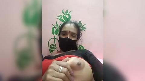 Media: A low-quality video of a South Asian woman with medium skin tone, black hair, and a black mask, exposing her breast with a red bra and ring, against a green wall.