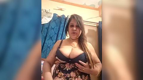 Media: Video of a plus-size woman with long brown hair, wearing a dark floral dress, lifting her breasts in a bathroom with blue shower curtain and floral wallpaper.