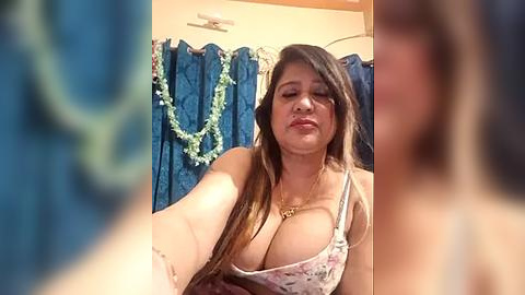 Media: Video of a young, curvy Latina woman with long brown hair, wearing a low-cut floral top, taking a selfie in a bathroom with blue curtains and a green necklace.