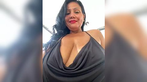 Media: Video of a smiling, voluptuous Black woman with long black hair, wearing a low-cut black dress that reveals ample cleavage. The background is blurry, focusing attention on her.