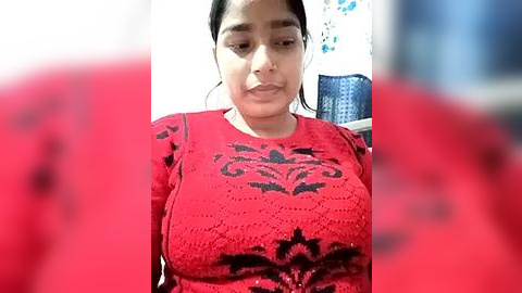 Media: Video of a South Asian woman with medium skin tone, wearing a red knitted sweater with black floral patterns, seated indoors against a blurred background.