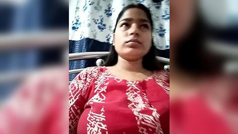 Media: Video of a South Asian woman with medium skin tone, long black hair, and a serious expression, wearing a red patterned blouse, seated in a hospital bed with white and blue curtains in the background.