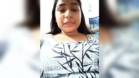 Media: Video of a young South Asian woman with medium skin tone, long black hair, and a serene expression, wearing a white blouse with a black leaf pattern, indoors with a blurred background.