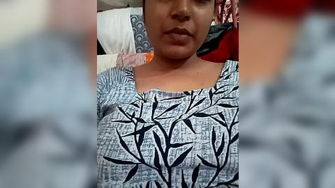 Media: A video of a woman with medium brown skin, wearing a black and white leaf-patterned top, sitting against a background of pillows and blankets.