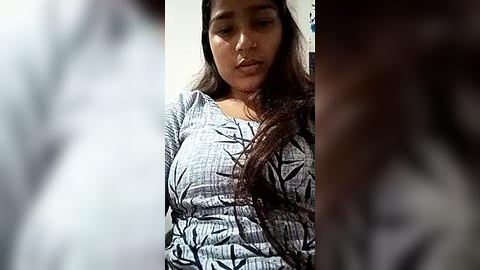 Media: Video of a South Asian woman with long, dark hair, wearing a patterned top, looking introspective in a dimly lit room.