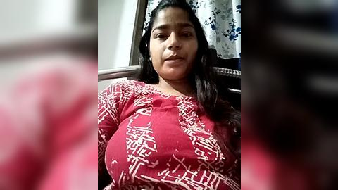 Media: A video of a South Asian woman with medium skin tone, long black hair, and a confident expression, wearing a red top with white geometric patterns. The background features a curtain with blue and white floral designs.