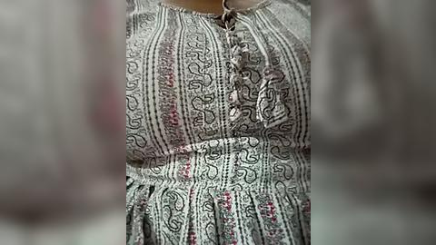 Media: A close-up video of a woman's upper torso wearing a long, intricately patterned, multi-colored, flowy dress with a high neckline and a small, decorative pocket on the chest. The background is blurred, emphasizing the dress's detailed embroidery.