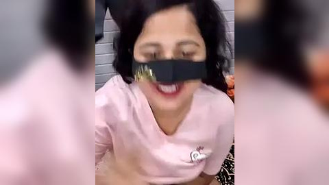 Media: Video of a woman with light skin and dark hair, wearing a pink top, black blindfold, and gag, kneeling on a floor with a blurred background.