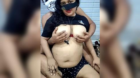 Media: A video captures a plus-sized woman with medium skin tone and curly black hair, wearing a black mask, lifting her large breasts with black polka-dot panties.