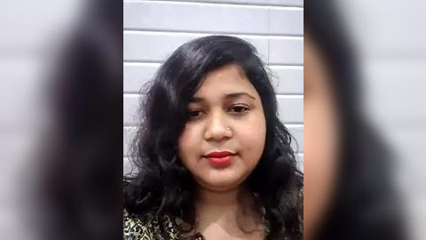 Media: Video of a young woman with medium skin tone, wavy black hair, and red lipstick, smiling against a white tiled wall. She wears a patterned blouse.