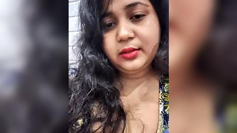 Media: Video of a young South Asian woman with medium-dark skin tone, curly black hair, and red lipstick. She wears a floral-patterned top. The background is blurred, focusing on her face.