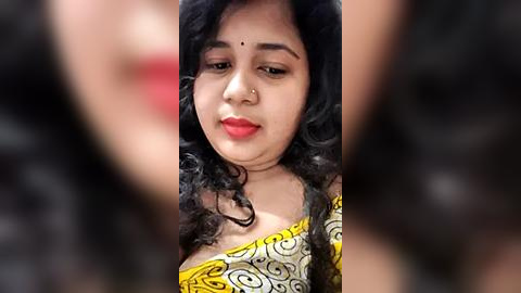 Media: Video of a young South Asian woman with fair skin, wearing a yellow saree with a black floral pattern, black curly hair, and a small nose ring. Background is blurred, focusing on her face.