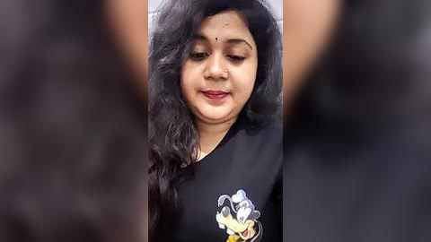 Media: Video of a South Asian woman with long, wavy black hair, wearing a black shirt with a cartoon character graphic. She has a slight smile, with a dot on her forehead, and is standing outdoors against a blurred background.