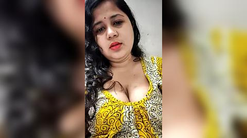 Media: A video of a young woman with fair skin, long black hair, and a red bindi. She wears a yellow and white floral blouse, revealing ample cleavage. Background is blurry.