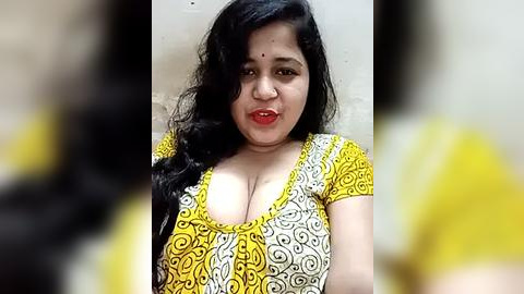 Media: Video of a South Asian woman with long, wavy black hair and fair skin, wearing a low-cut yellow and white patterned blouse revealing ample cleavage. The background is blurred, focusing on her face.