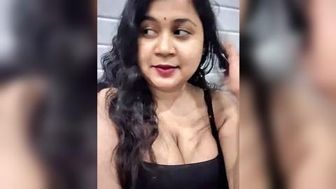 Media: A video of a middle-aged South Asian woman with long, wavy black hair, wearing a black tank top, smiling in a bathroom with tiled walls.