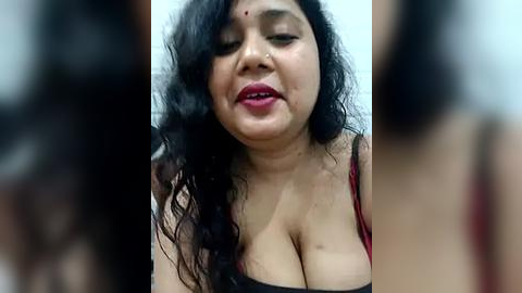 Media: A video of an Indian woman with medium brown skin, curly black hair, and a red dot on her forehead, wearing a red bra, showing cleavage, smiling, with blurred background.