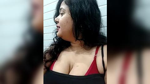 Media: A video of a plus-sized woman with long, wavy black hair, wearing a red bra and black tank top, standing against a tiled bathroom wall.