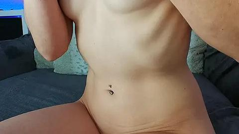 Media: Video of a nude, light-skinned individual with a slender physique, showcasing a flat stomach, pierced navel, and partial arm. They sit on a dark gray couch, with a blue-tinted background.