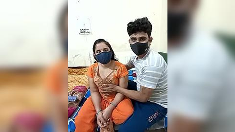 Media: Video of a couple sitting on a blue mat in a room with white walls and a brown carpet. The woman, wearing an orange sari and blue mask, looks distressed, while the man, in a white striped shirt and blue mask, appears calm.