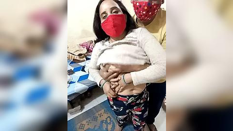 Media: Video of a pregnant woman with a red mask and white top, standing in a cluttered room with a bed, a child, and another person in the background.