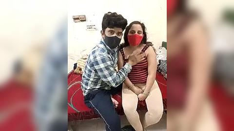 Media: Video of a man in a plaid shirt and mask, holding a woman in a red mask and striped top, sitting on a bed in a dimly lit room.