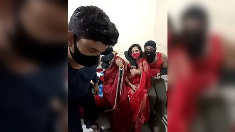 Media: Video: Three masked men in red outfits, one holding a gun, forcing a woman in red attire to sit on a couch; background blurred.