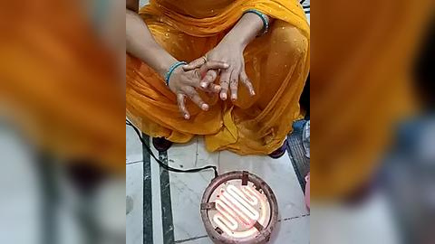 Media: Video of a person sitting on the floor, hands clasped, wearing an orange sari. A brass bell lies on the tiled floor in the background.