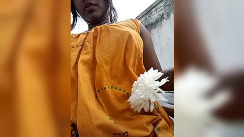 Media: Video of a South Asian woman with medium-brown skin, wearing a yellow sari, holding a white flower. Background shows a white wall and orange fabric.