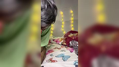 Media: Video of a woman with shoulder-length gray hair, wearing a green shirt, standing beside a bed with a colorful, butterfly-patterned duvet, lit by yellow fairy lights.