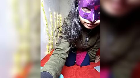 Media: A video of a woman with long black hair, wearing a purple mask and green jacket, sitting on a red carpet with yellow string lights in the background.