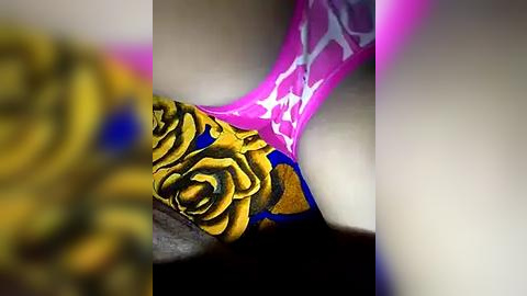 Media: Video of a close-up tattoo on a person's arm, featuring a detailed, vibrant rose design in gold and black ink, with a glowing pink flame-like pattern overlaid. The image is blurred and grainy, likely taken with a low-quality camera.