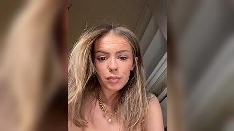 Media: A video of a young, fair-skinned woman with shoulder-length, straight blonde hair, wearing a gold necklace, making a pouty face. The background is blurred, showing a white door and beige wall.