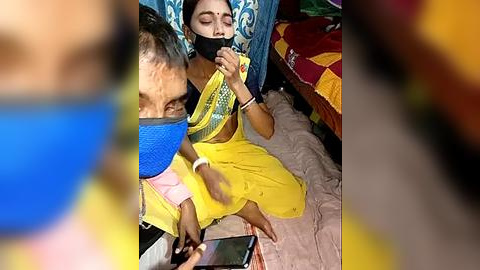 Media: Video of a woman in a yellow sari, gagged, sitting on a bed with a blue curtain. Another person, blurred, is holding a phone. Background includes a colorful bedspread and a blue cushion.