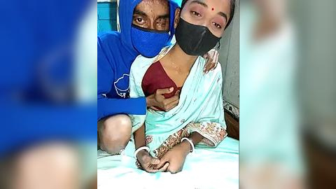 Media: Video of a young woman with a black face mask, wearing a blue saree and a red blouse, being held by a man in a blue shirt and a mask.