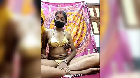 Media: A video of a woman meditating, wearing a gold bikini and a black face mask, surrounded by vibrant, patterned tapestries in a dimly lit room.