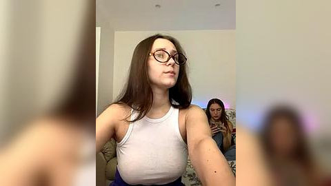 Media: A video of two women in a living room. The woman in the foreground has long brown hair, wears glasses, and a white tank top, while the woman in the background, blurred, looks down at her phone.