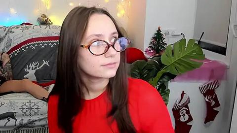 Media: Video of a fair-skinned woman with long brown hair and glasses, wearing a red sweater, sitting in a cozy, festive room with holiday decorations, including a Christmas tree and hanging lights.