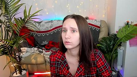 Media: Video of a young Caucasian woman with long brown hair, wearing a red plaid shirt, sitting indoors on a gray couch with red and white patterned blanket, surrounded by potted plants and fairy lights.