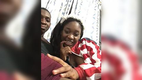 Media: Video of a smiling young Black woman in a red and white striped hoodie, sitting on the lap of a man in a dark shirt, both wearing white headbands. The background features a white curtain with a subtle floral pattern.