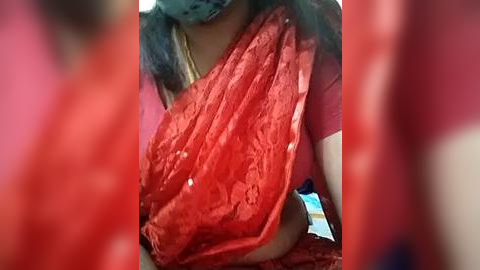 Media: Video of a person with dark skin wearing a red, floral-patterned saree, partially covering their chest. The background is blurred, featuring muted colors. The saree's texture appears delicate and lace-like.