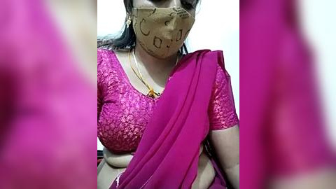 Media: Video of an Indian woman in a bright pink saree and beige mask with \"OM\" pattern, sitting indoors, blurred background.