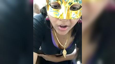 Media: Video of a young woman in a gold Venetian mask, purple top, and gold necklace, leaning forward in a crowd, her expression intense and lips slightly parted.